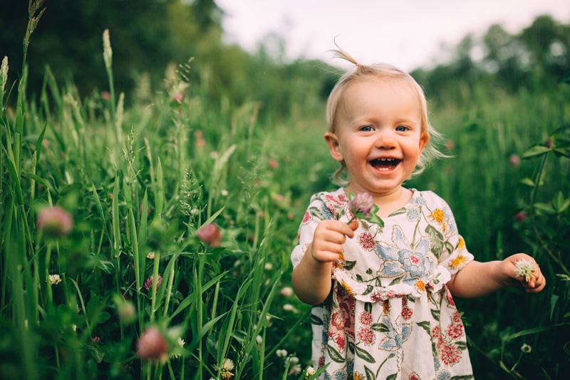 Milestone Photography, a young toddler girl laughs as she walks through tall grass with a flower in her hand