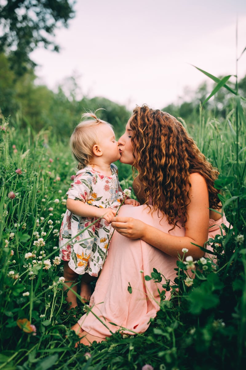 Motherhood Photography, a young mother kisses her baby girl, they sit in the a flowery grassy meadow