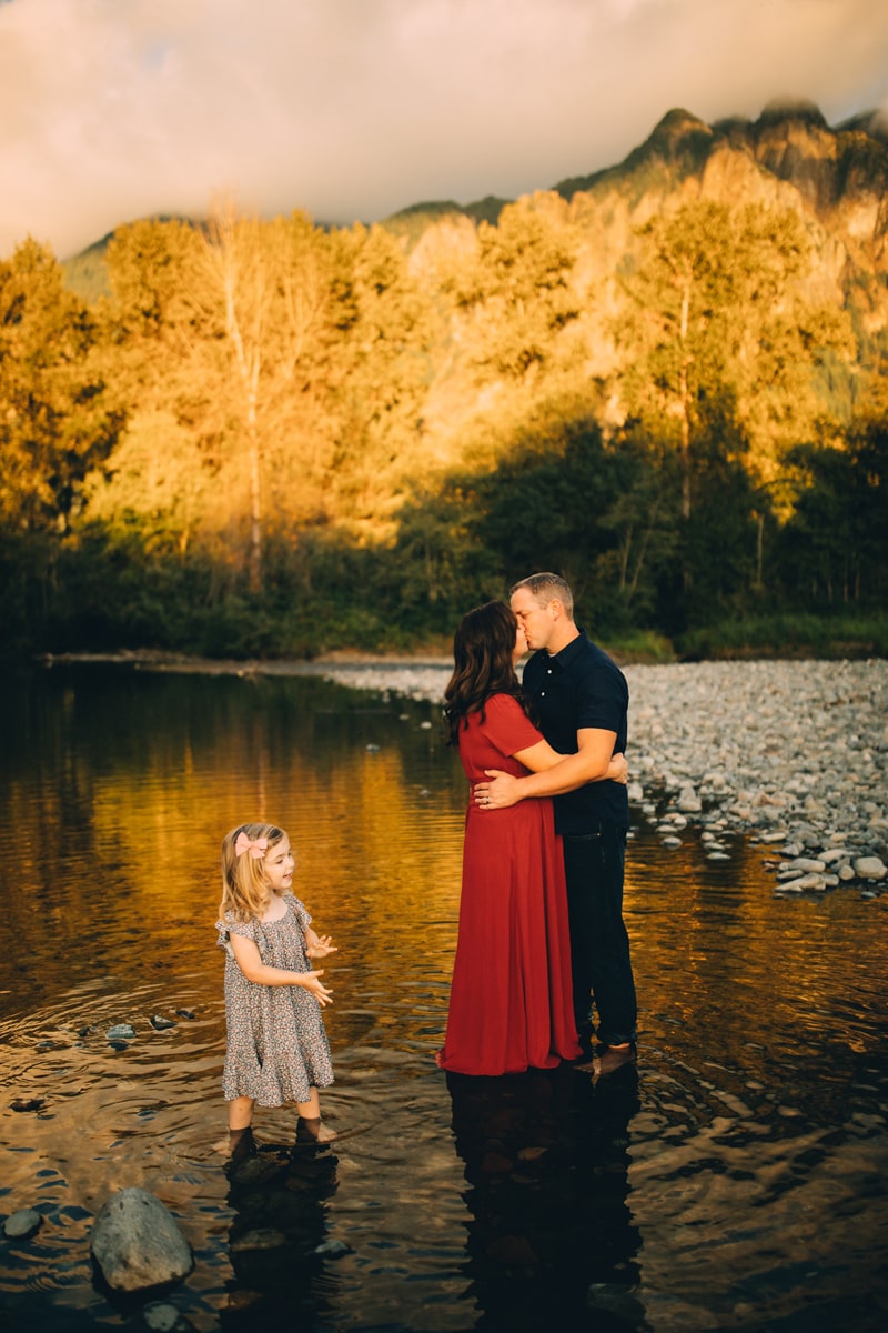 Family Photographer - husband and wife kiss in quiet stream, toddler daughter smiles next to them